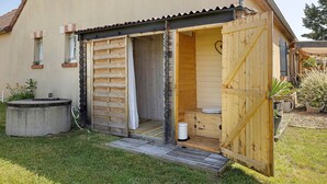 Une salle de bain extérieure en bois, équipée d’une douche, de toilettes et d’un petit bac à fleurs en béton.