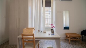 Una habitación con escritorio de madera, una silla, una mesita, un taburete de mimbre, una ventana con cortinas y un aire acondicionado instalado en la pared.