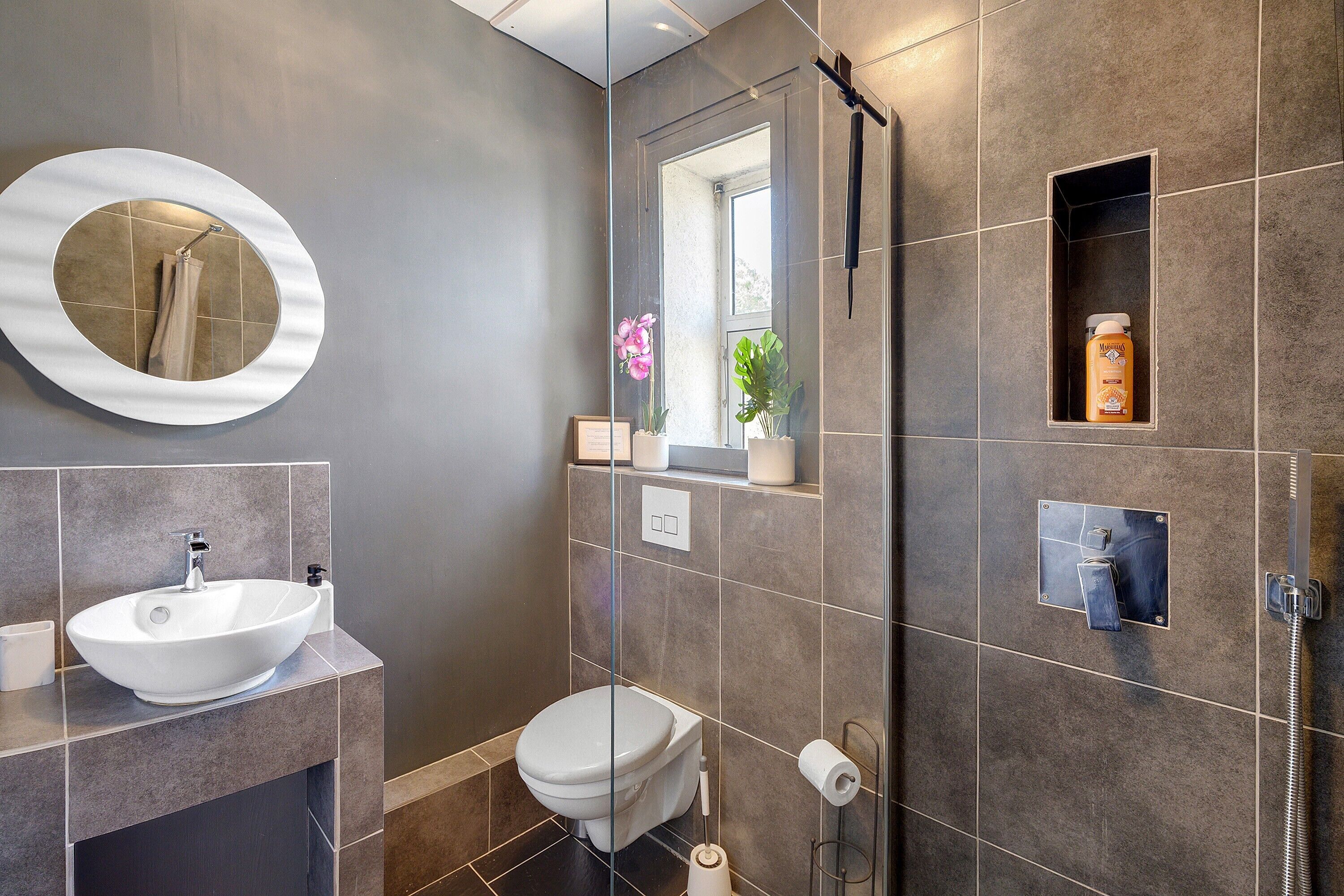 Et moderne badeværelse med et rundt spejl, en hvid vask, toilet, brusebad og et lille vindue med planter.
