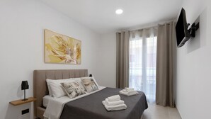 A neatly made bed with a dark bedspread, a bedside table with a lamp, a wall-mounted TV, and a large flower painting. - Room 'Camera 3 - P1 Con Accesso Disabili' with Balcony, Wi-Fi and Air Conditioning (Balestrate)