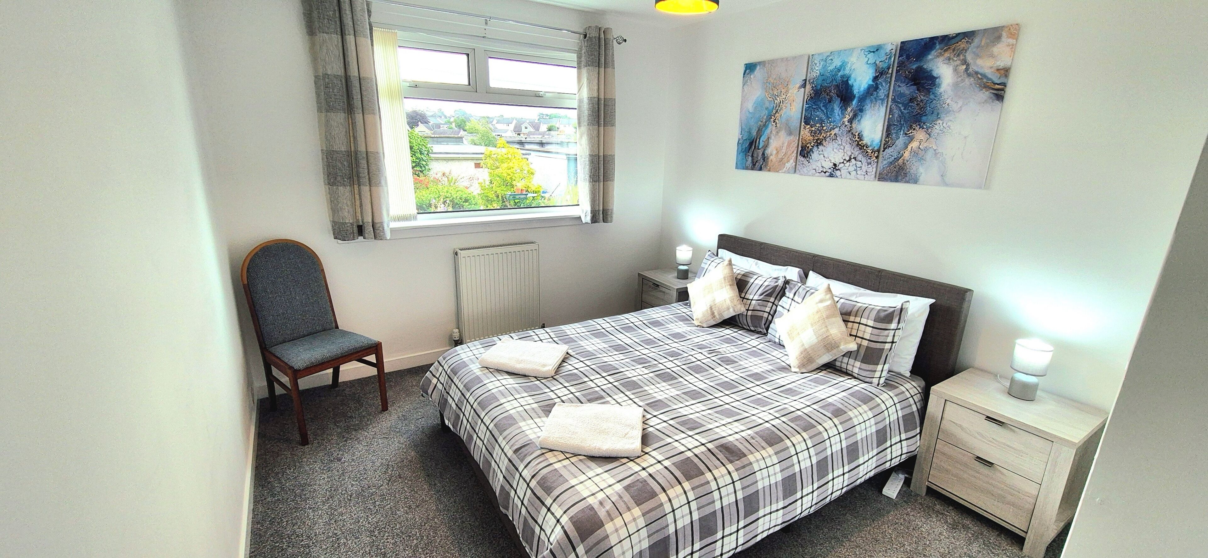 A bedroom with a bed, a chair, a nightstand, a radiator, a window with curtains, and a painting on the wall.