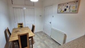 Un comedor con una mesa de madera, sillas y un florero con flores. Hay puertas blancas y un radiador en la pared.