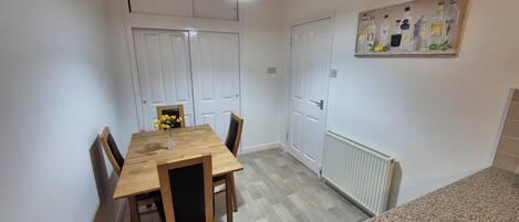 Un comedor con una mesa de madera, sillas y un florero con flores. Hay puertas blancas y un radiador en la pared.