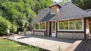 Een huis met een gevel van natuursteen, een houten terras en een groot raam met uitzicht op een bebost gebied.