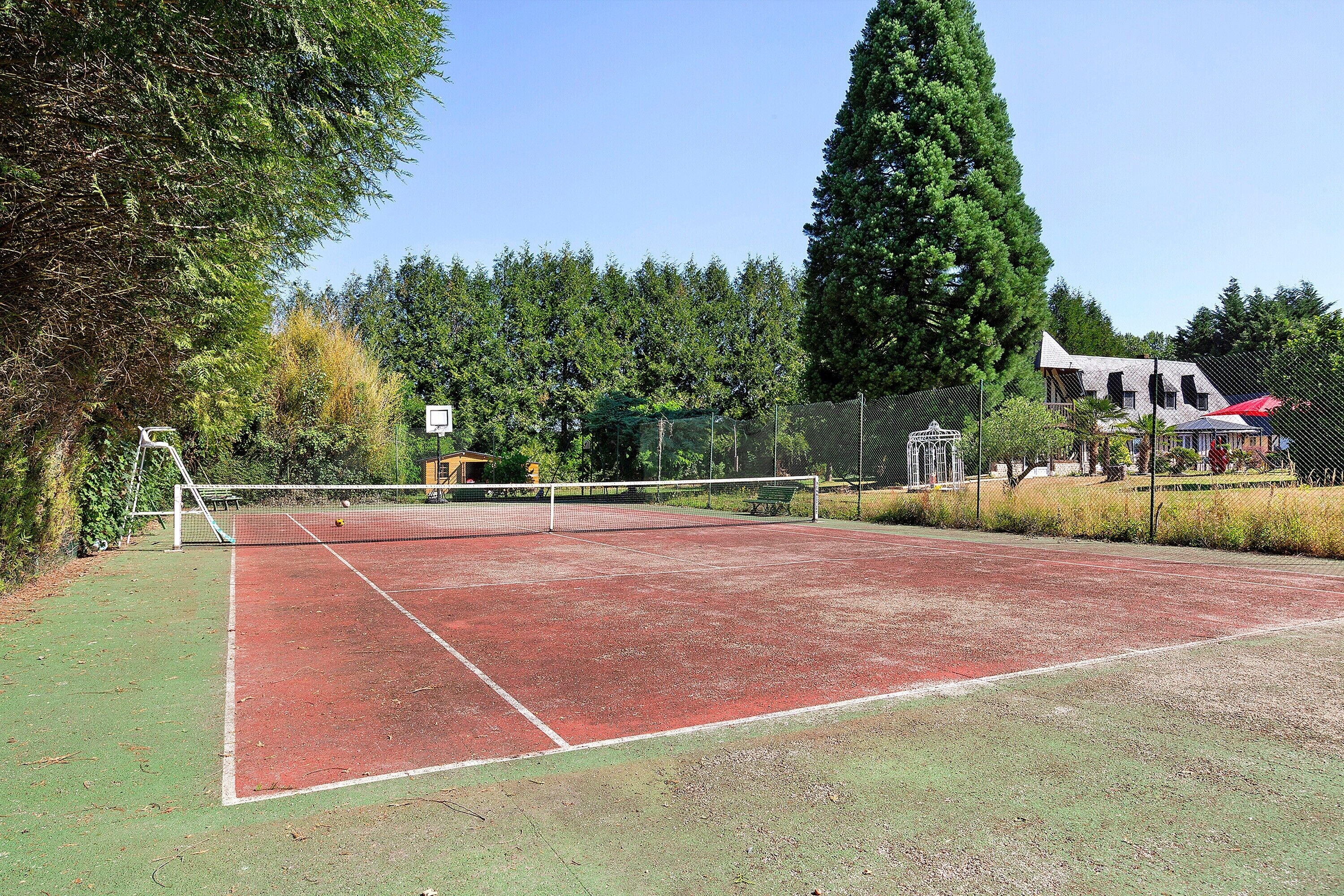 Un court de tennis avec un filet, entouré d’arbres et bordé d’un bâtiment dont le toit est rouge.