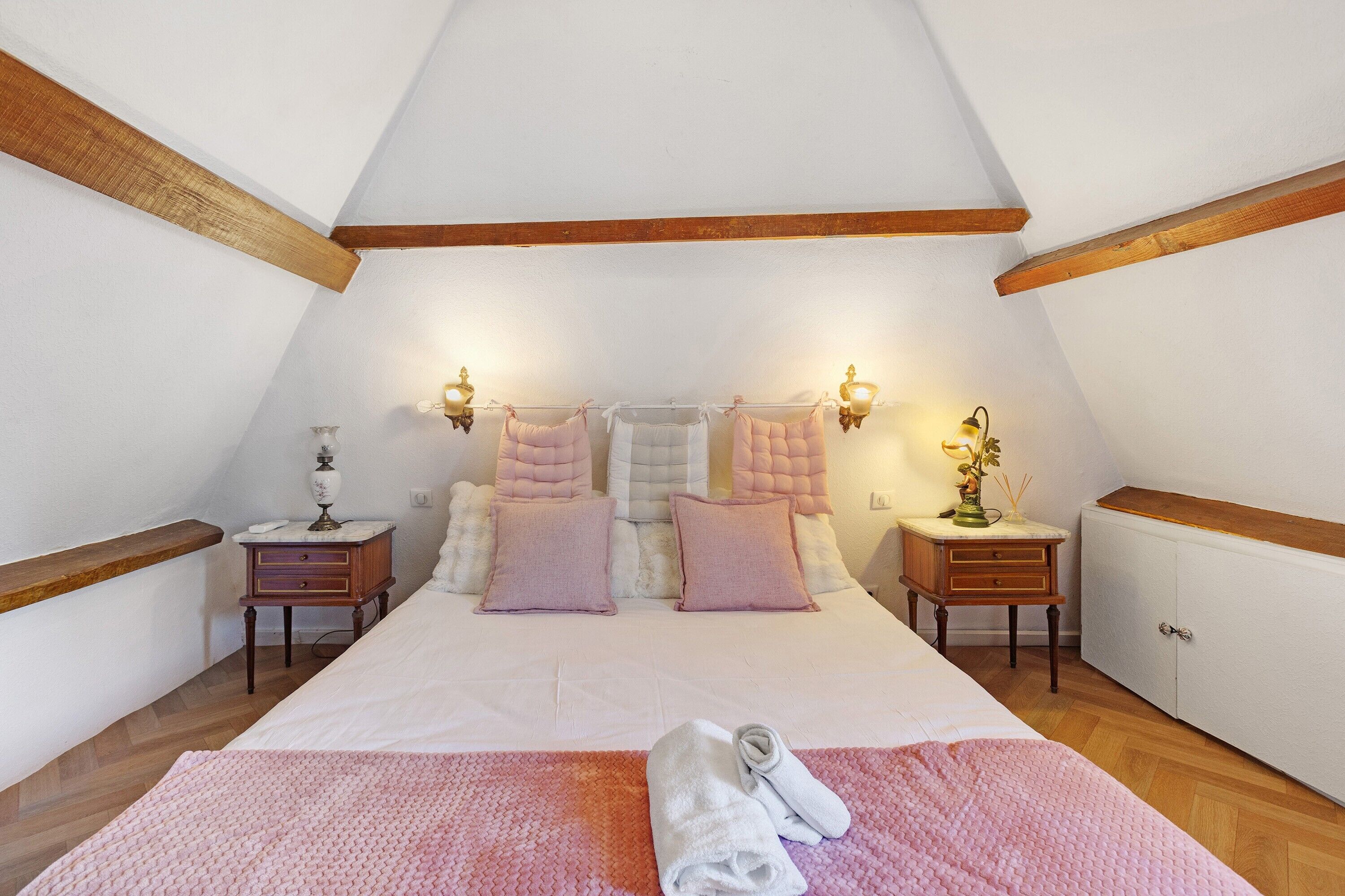 Un dormitorio con techo inclinado, vigas de madera, una cama con varias almohadas, dos mesitas de noche con lámparas y un radiador.