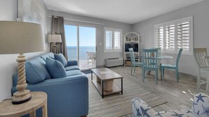 En rummelig stue med en blå sofa, et sofabord og et spiseområde med blå stole. Der er en balkon med havudsigt.