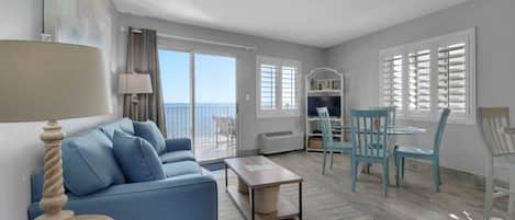 A spacious living room with a blue sofa, a coffee table, and a dining area with blue chairs. There is a balcony with a view of the sea.