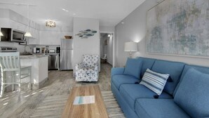 En moderne stue med en blå sofa, et træsofabord og et køkkenområde med hvide skabe og stålemmer.
