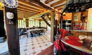 Un interior rústico con vigas de madera a la vista, una zona de comedor con mesa y sillas, y una cocina con electrodomésticos modernos.