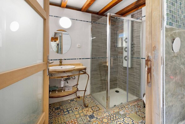 Un baño con ducha envuelta en vidrio, lavamanos con tocador y baldosas de piso estampadas.