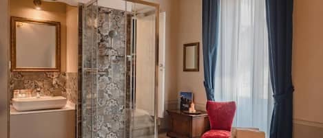 Camera d'albergo con cabina doccia in vetro, un letto con una poltrona rossa, un comodino con una lampada e un'ampia finestra con tende.