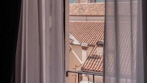 A window with sheer curtains, a view of a building with a tower, and a tiled roof. - Palazzo dei Pavoni Relais di Charme (L'Aquila)