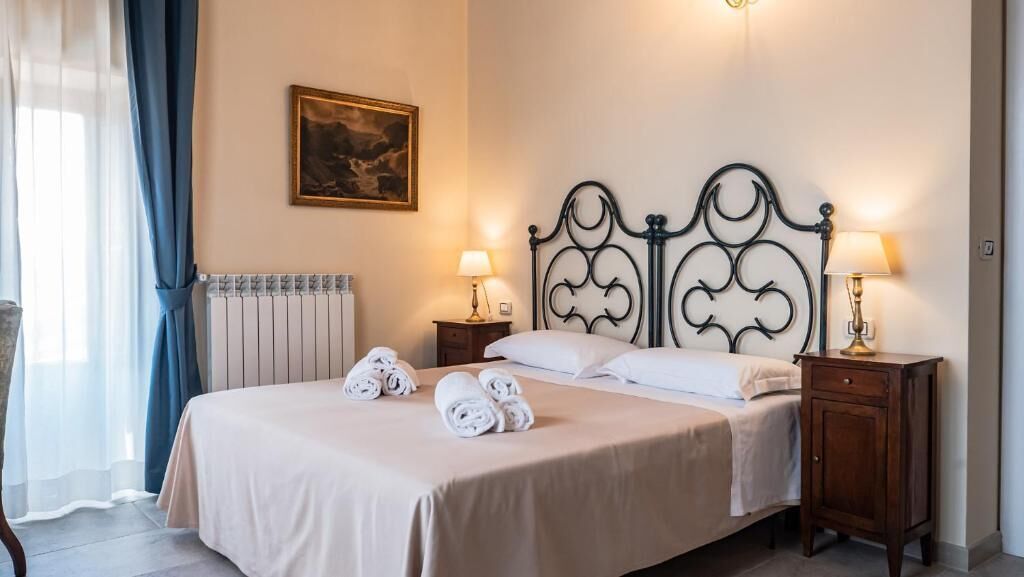 Una cama bien hecha con sábanas blancas y toallas enrolladas, cabecera de metal clásico, lámparas de noche y un cuadro enmarcado en la pared.