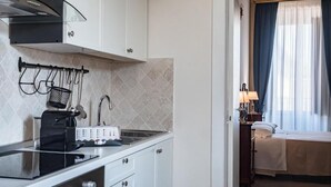 A compact kitchen with a toaster, kettle, and a small sink. Adjacent is a bedroom with a bed, nightstand, and a lamp. - Palazzo dei Pavoni Relais di Charme (L'Aquila)