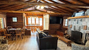 A log cabin interior with a wooden dining table, chairs, a sofa, a fireplace, and a television.