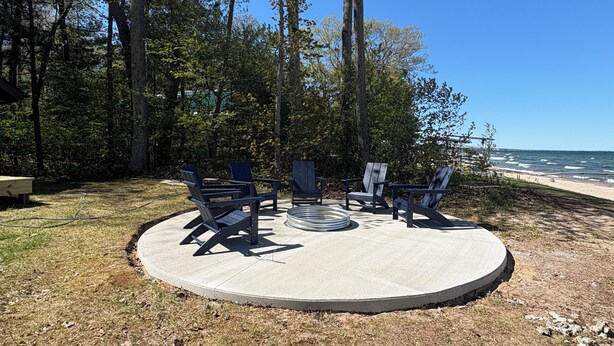 A circular outdoor seating area with Adirondack chairs and a fire pit, surrounded by trees and a view of the beach.