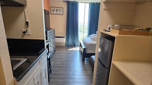 Cozinha compacta com geladeira, fogão e pia. Uma cama é visível ao fundo, com uma janela e cortinas.