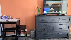 Quarto com televisão exibindo um anúncio da Louis Vuitton, uma escrivaninha com cadeira, um cesto de lixo e um armário com gavetas.