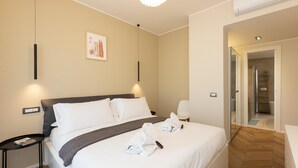 A hotel room with a bed, bedside tables, a mirror, a painting on the wall, and a hallway leading to a bathroom. - Rental in Rome Aurelia Sunset Penthouse (Rome)
