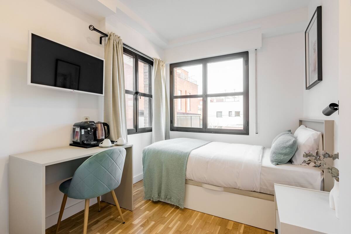 A modern bedroom with a bed, a desk, and a chair. There is a window with curtains, a coffee maker on the desk, and a picture frame on the wall.