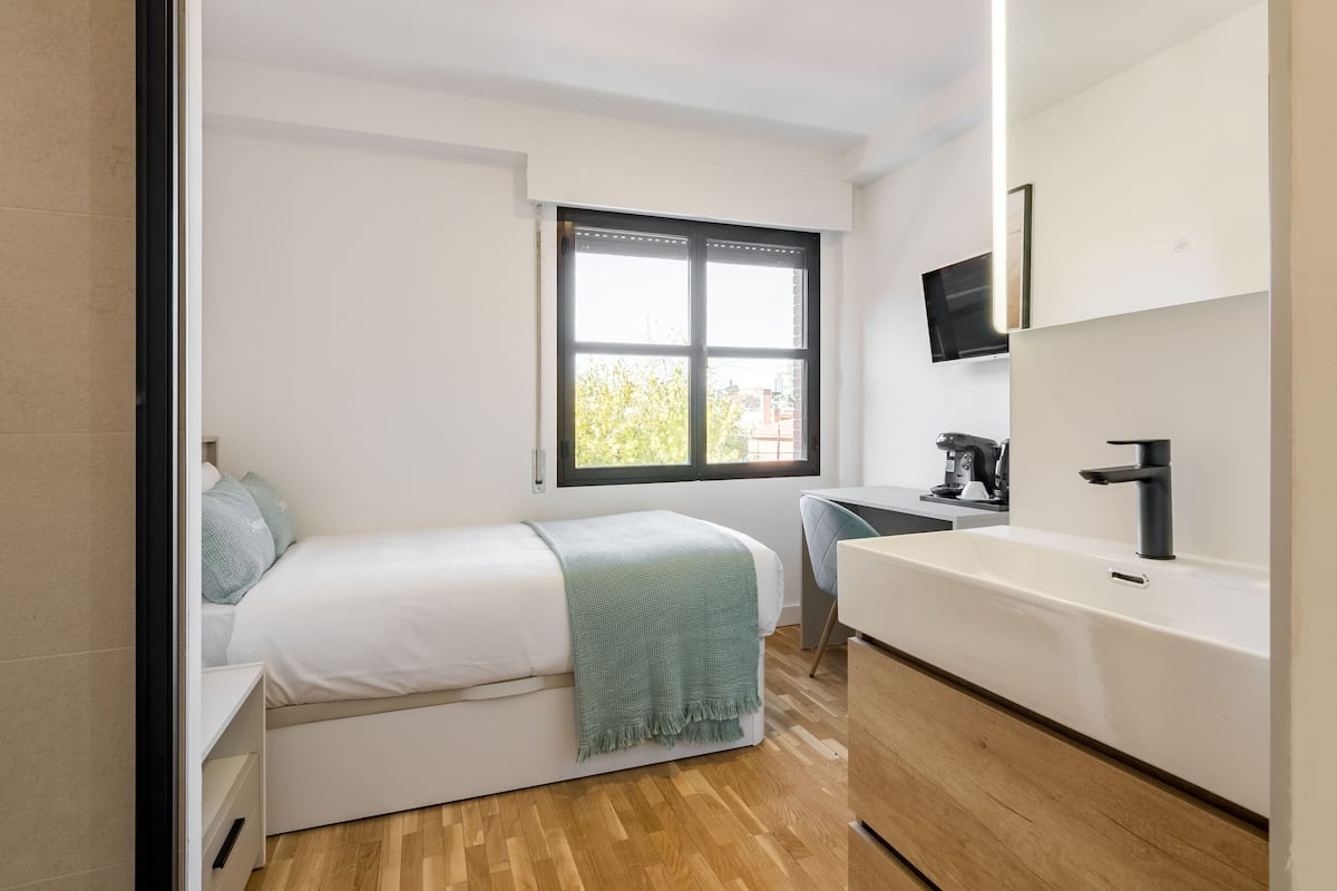 A modern bedroom with a bed, a desk, and a bathroom with a sink and mirror.