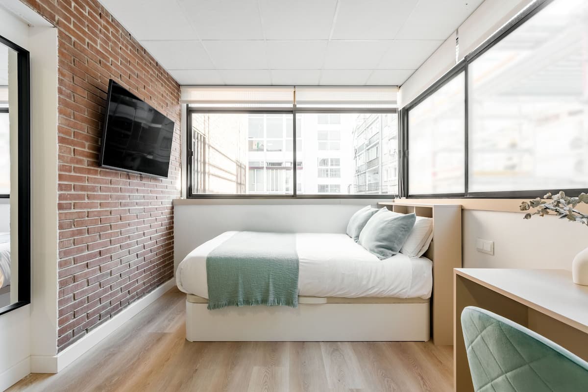 A modern bedroom with a brick wall, a bed with a green throw, a desk, and a TV mounted on the wall.