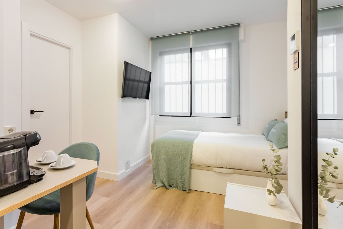 A minimalist bedroom with a bed, a TV mounted on the wall, a coffee maker, and a small table with cups.