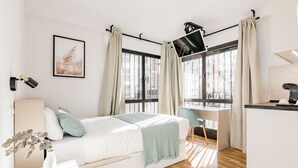 A modern bedroom with a bed, desk, and chair, large windows with curtains, and a city view. - Square Rooms by Charming (Madrid)
