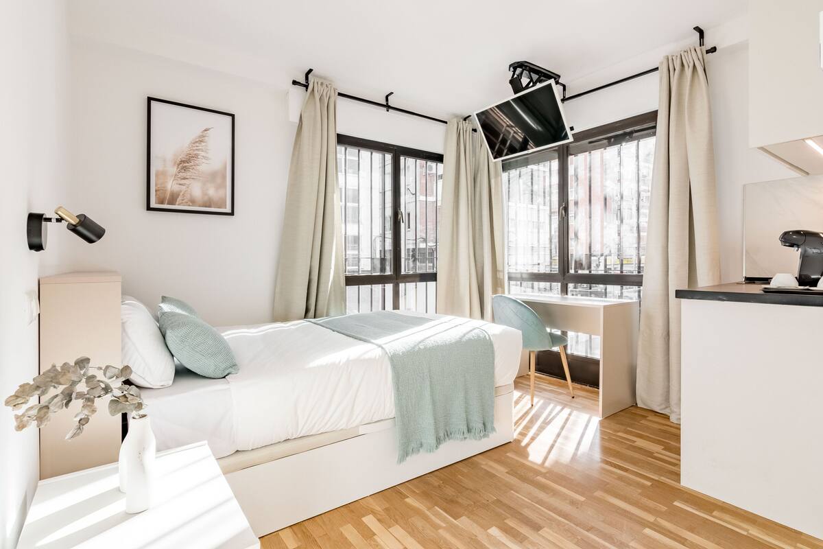 A modern bedroom with a bed, desk, and chair, large windows with curtains, and a city view.