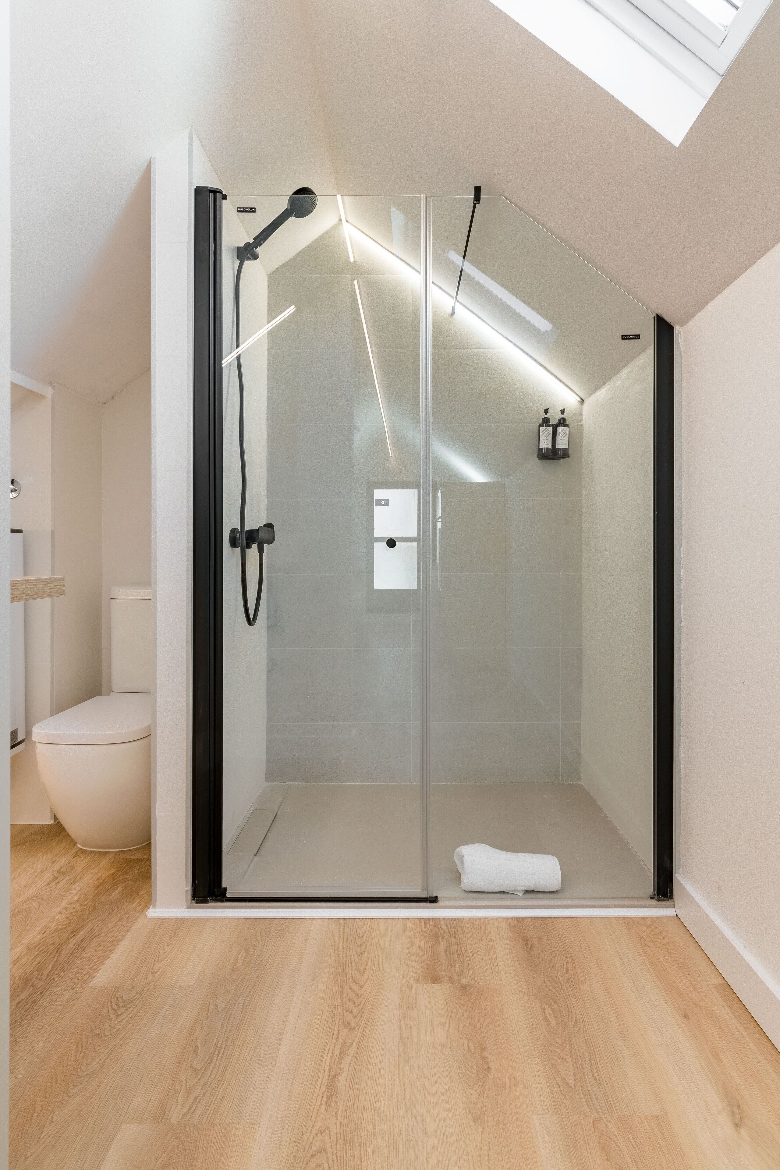 Un baño moderno con mampara de vidrio, inodoro blanco y suelo de madera.