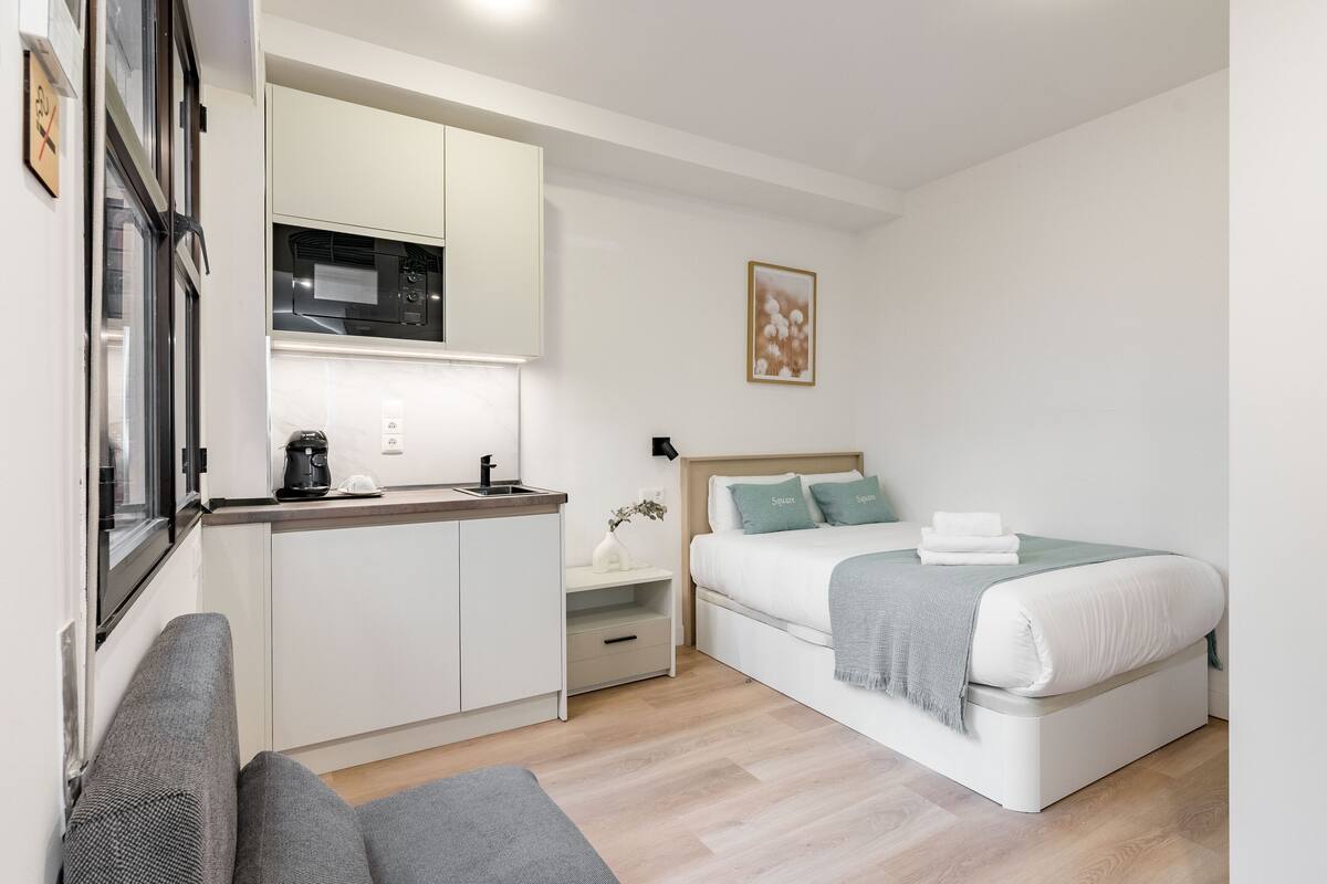 A compact room with a kitchenette, a bed with pillows, a bedside table, and a gray sofa.