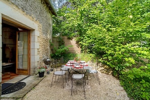A stone building with a wooden door, a gravel patio, a table with chairs, and a garden with green foliage.