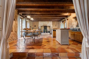 A rustic kitchen with a stone wall, wooden beams, a tiled floor, and a dining area with a table and chairs.