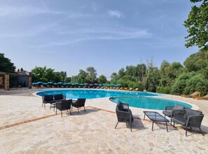 Zona de piscina con sillas de descanso y sombrillas, rodeada de árboles.