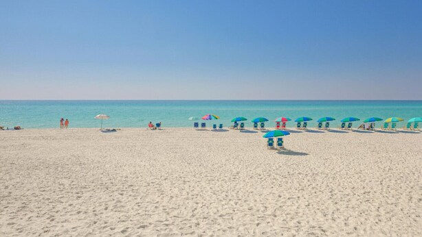 Una playa con arena blanca y cielo azul despejado. Hay sombrillas coloridas y sillas de descanso a lo largo de la orilla.