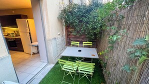 Een klein terras buiten met een tafel en stoelen, een koelkast en een keuken die zichtbaar is door een open deur.