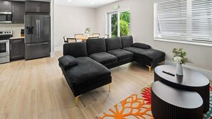 Un salon moderne comprenant un canapé d’angle, un coin repas et une cuisine équipée d’appareils électroménagers en acier inoxydable.