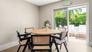 Une pièce avec une table ronde en bois et six chaises, un vase blanc rempli de fleurs, et une vue sur un jardin par une porte vitrée.