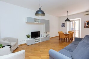 Una sala moderna con un sofá gris, un sofá blanco, un televisor sobre un mueble blanco y una zona de comedor con una mesa y sillas.