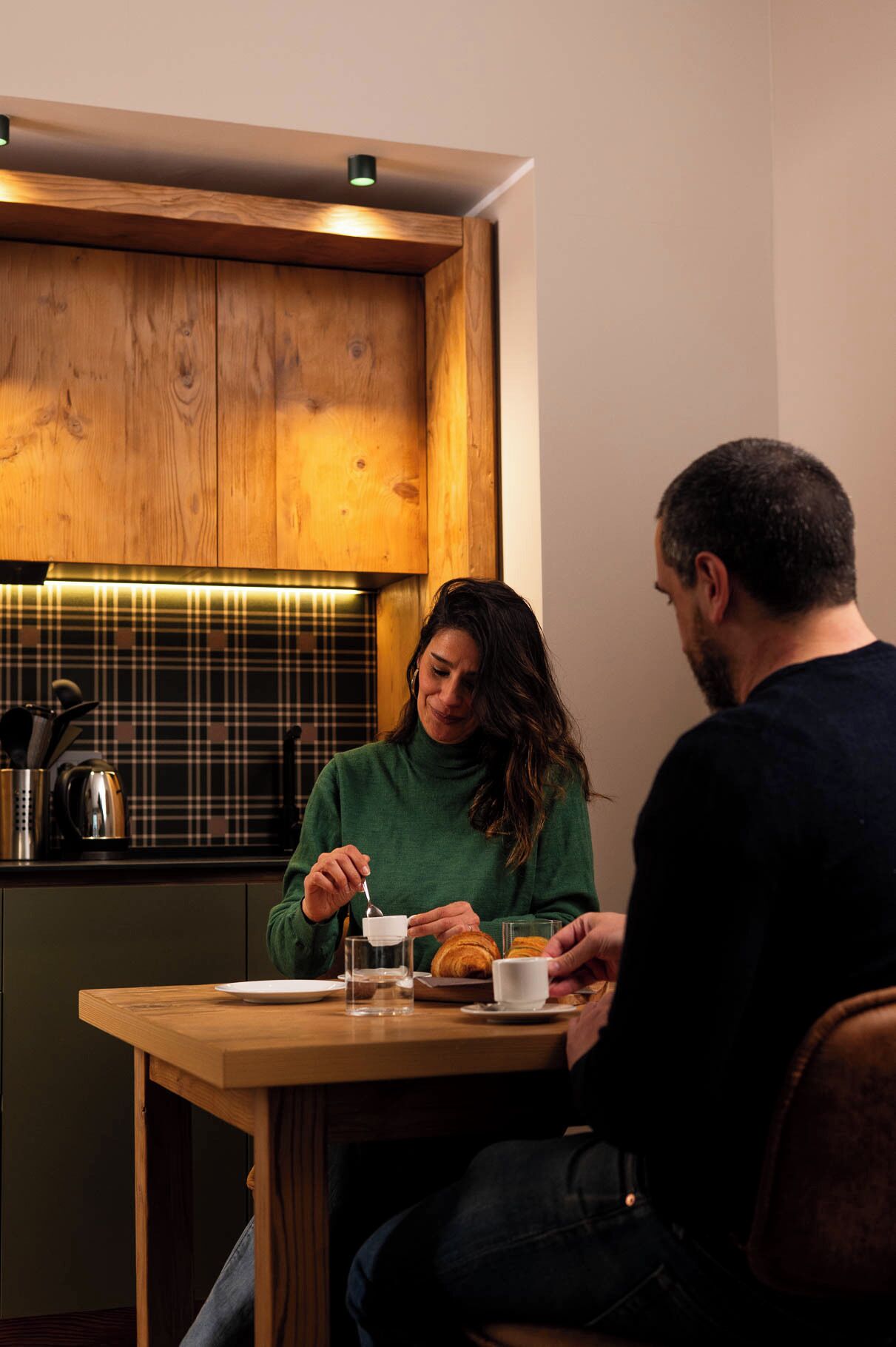 A cozy dining area with a wooden table and chairs, a plaid backsplash, and a modern kitchen in the background.