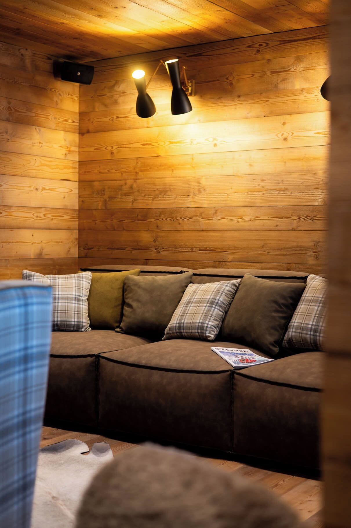 A cozy cabin interior with a wooden wall, a brown sofa with plaid cushions, and two pendant lights.