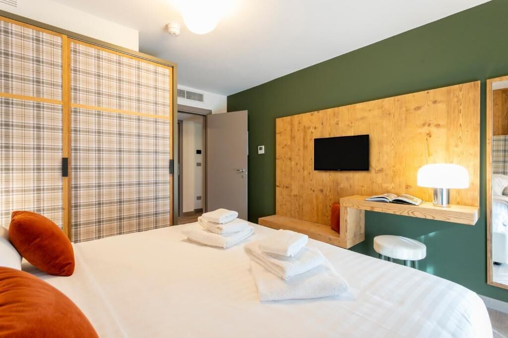 A hotel room with a bed, a wooden headboard, a TV, a bedside table with a lamp, and a wardrobe with a plaid pattern.