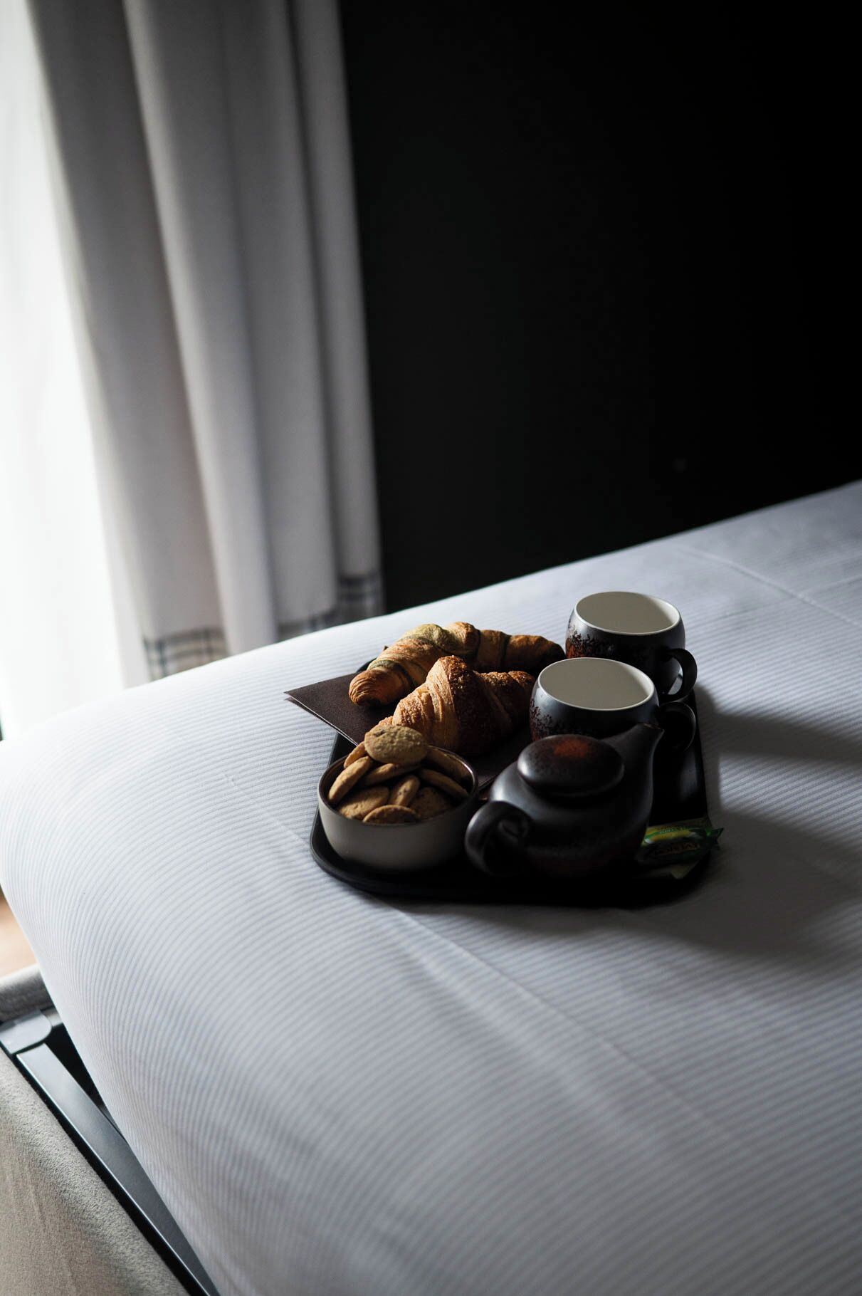 A neatly arranged breakfast setting on a bed with croissants, cookies, and two black cups, likely containing coffee or tea.