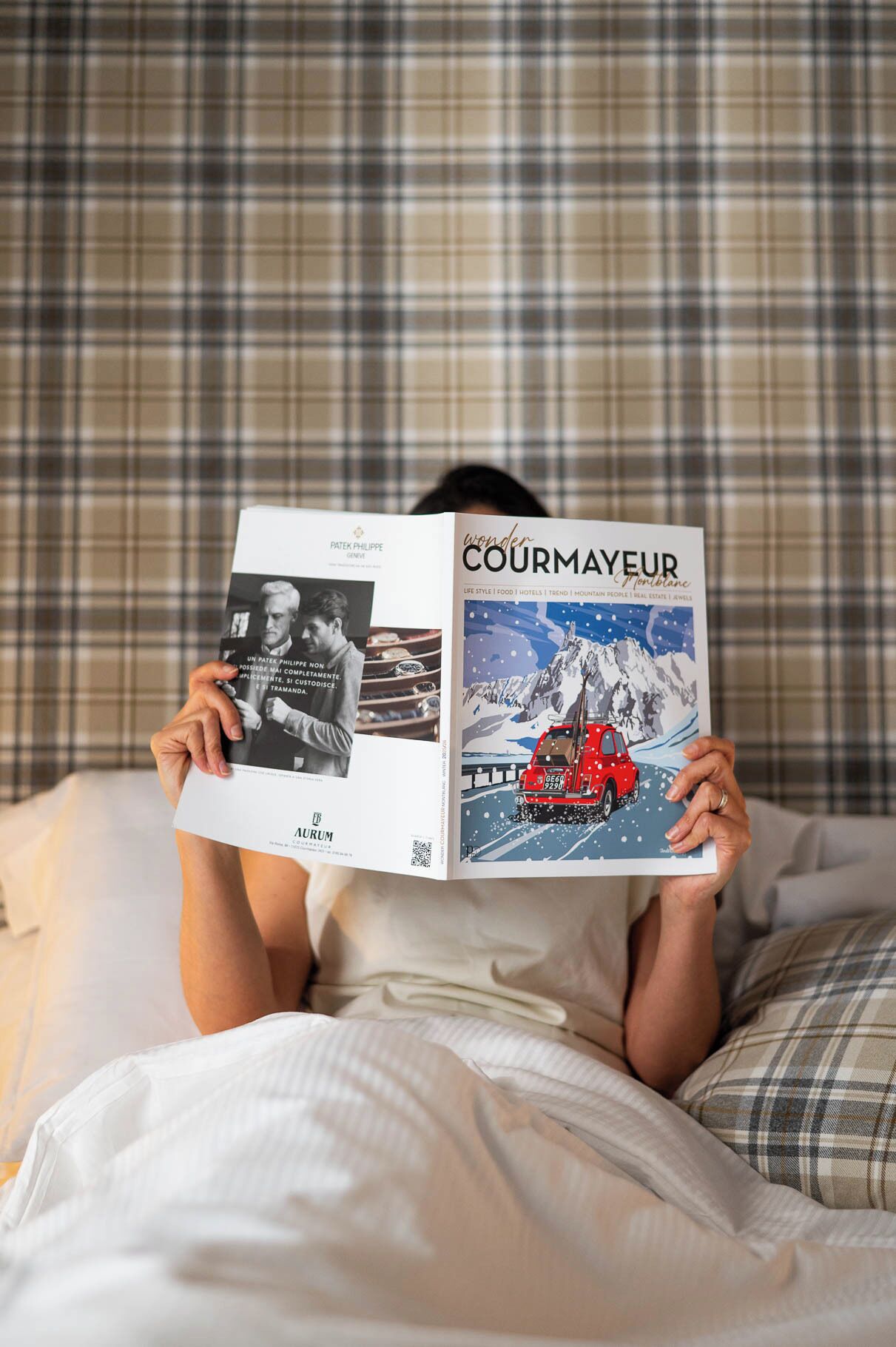 A person lying on a bed, holding a book titled 'COURMAEUR' with a winter scene on the cover.