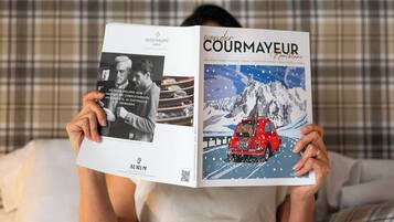 A person lying on a bed, holding a book titled 'COURMAEUR' with a winter scene on the cover.