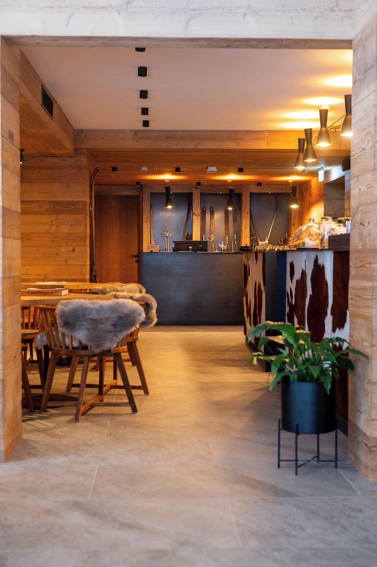 A cozy restaurant with wooden walls and a central bar area. There are wooden chairs with fur throws and a potted plant.