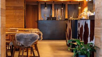 A cozy restaurant with wooden walls and a central bar area. There are wooden chairs with fur throws and a potted plant.