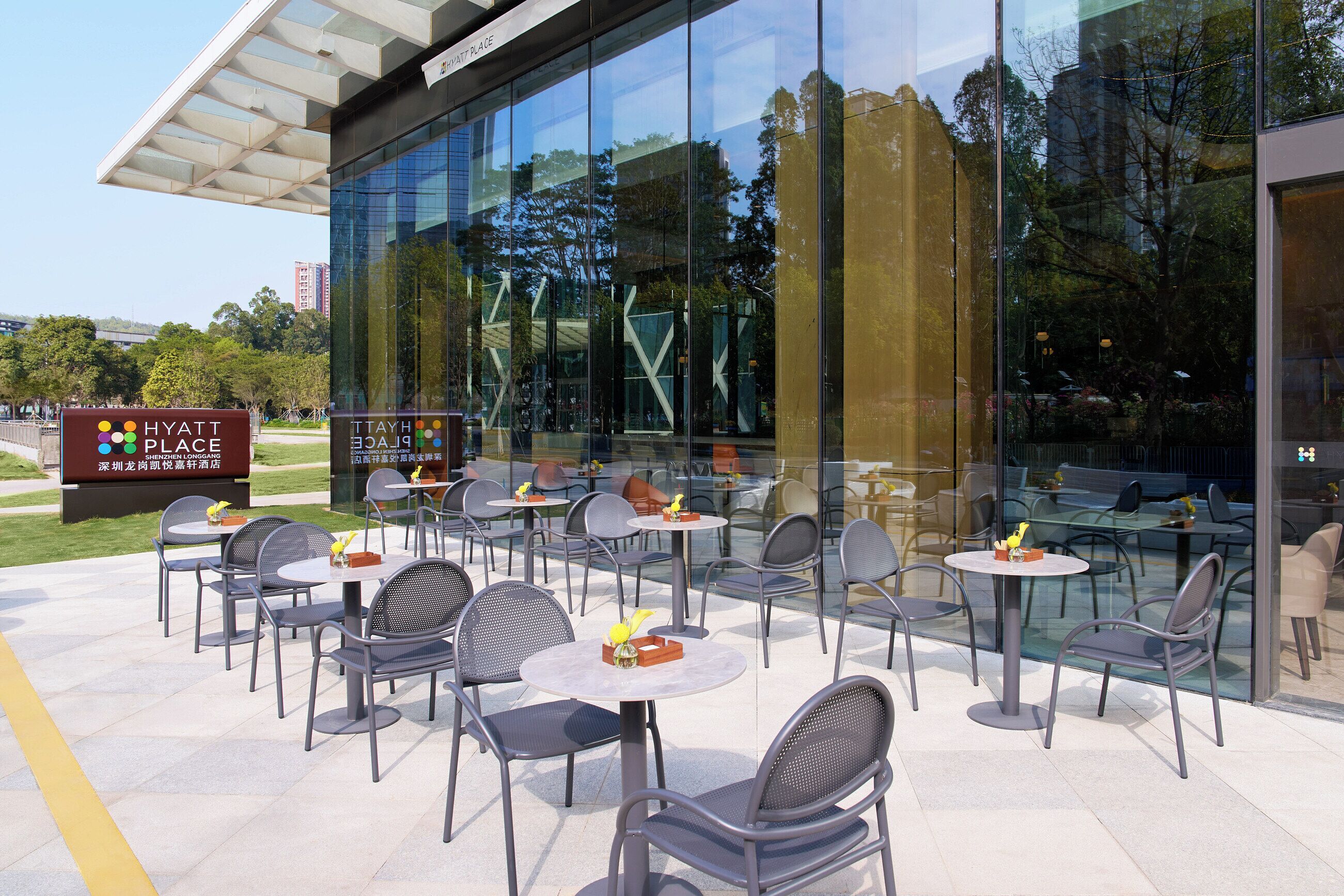 An outdoor seating area with tables and chairs, a sign for Hyatt Place, and a glass building reflecting the surroundings.
