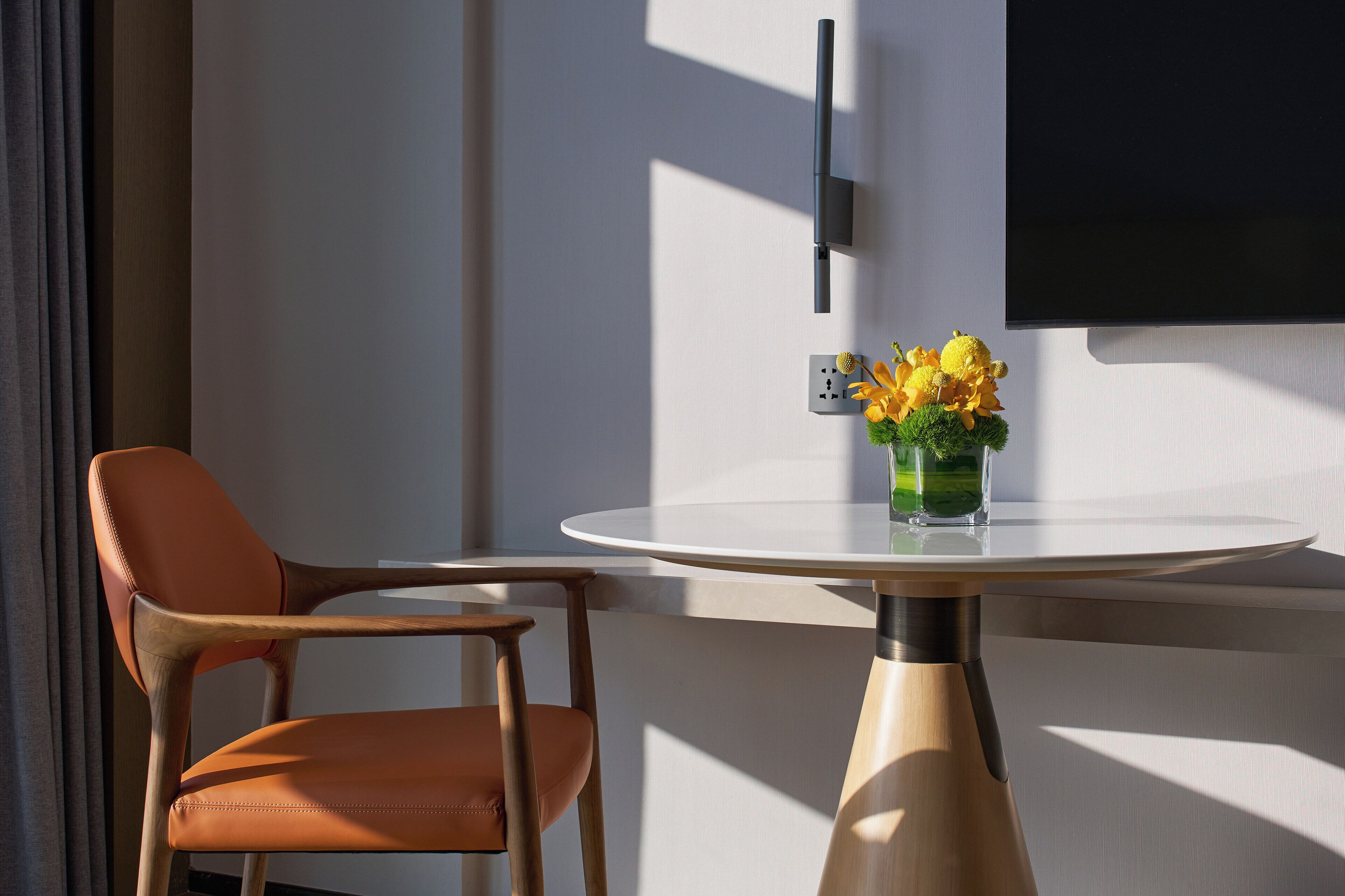 Una habitación moderna con una mesa redonda, una silla naranja individual, un televisor montado en la pared y un pequeño florero con flores.